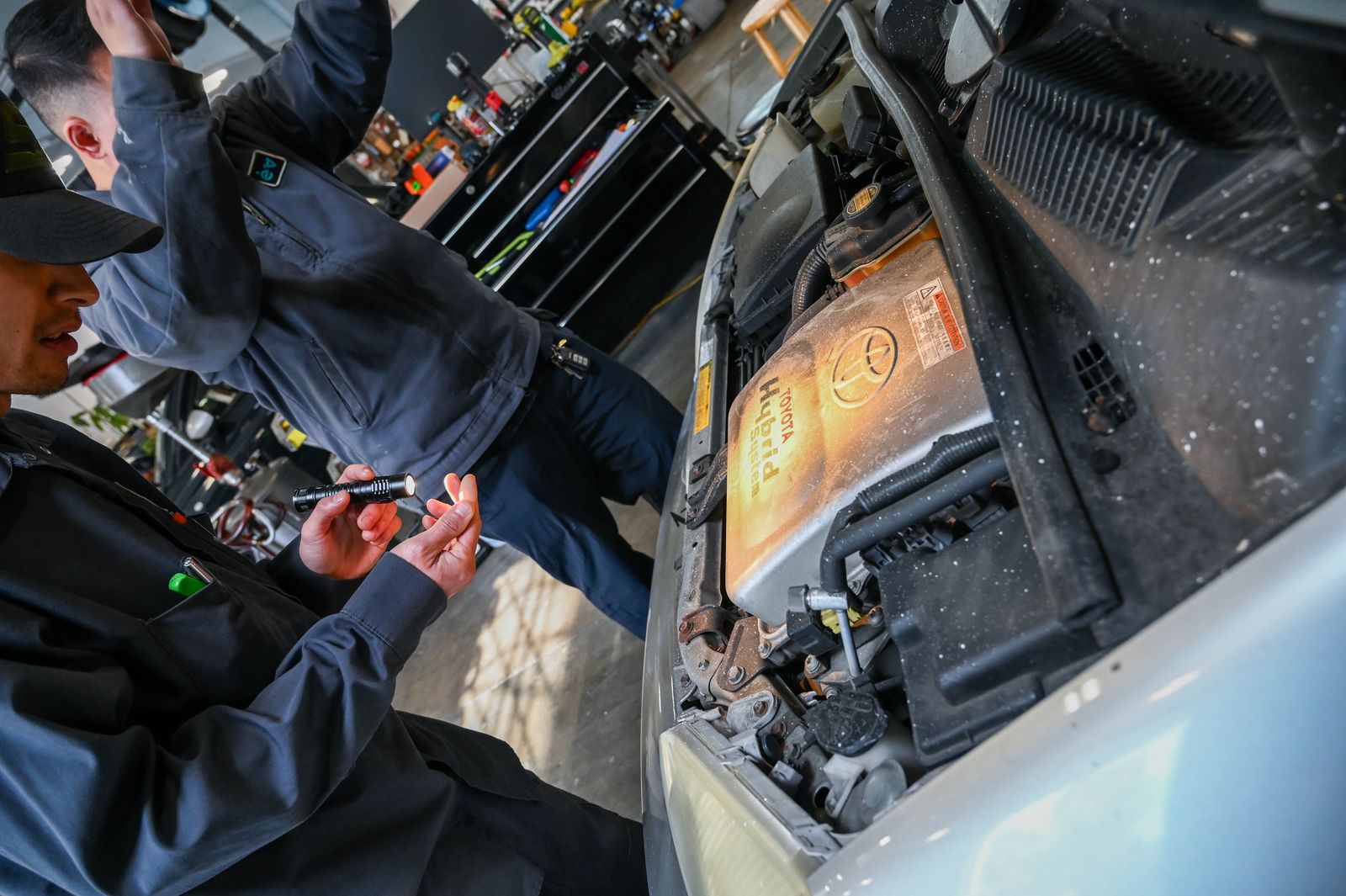 A technician's flashlight on a hybrid's engine bay, the orange high-voltage cover in view